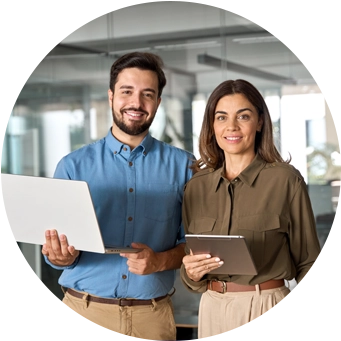 A man and a woman standing together in an office, holding a laptop and a tablet, ready to assist.